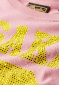 Rosa Baumwoll-T-Shirt mit großen, gelben, genoppten Buchstaben. Strukturierte, glänzende Verzierungen kontrastieren mit dem weichen Stoff.