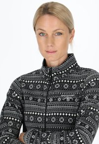 Blonde woman with blue eyes wearing a black and white patterned zip-up fleece jacket with arms crossed against a white background.