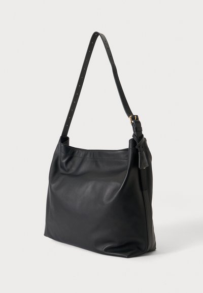 Sac à épaule en cuir noir avec sangle ajustable et boucle dorée posé sur un fond gris clair.