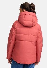 Veste rembourrée en corail avec une grande capuche arrondie. Elle présente un matelassage horizontal et une coupe décontractée, associée à un jean en denim bleu.