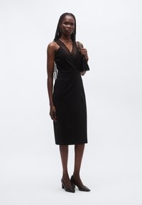 Must kleit sügava V-kaeluse ja tekstuurse pealiskihiga. Vormiv lõige, põlvpikkusega, küljetaskutega. Modell hoiab musta käekotti.