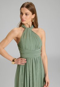 Robe verte à col halter en tissu plissé, taille cintrée et embellissements subtils. Un bracelet manchette doré complète le look.