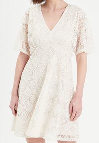 Robe en dentelle ivoire avec un décolleté en V, des manches courtes et une silhouette en A. Présente des motifs découpés complexes et une texture fluide.