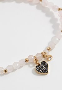 Un bracelet en perles de quartz rose avec des accents dorés, orné d'un charme en forme de cœur noir décoré de petites pierres et d'un détail en gemme claire.