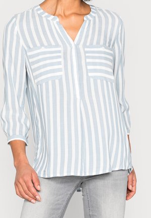Femme portant un chemisier à rayures verticales bleu clair et blanc avec deux poches poitrine à rayures horizontales et des manches trois-quarts.