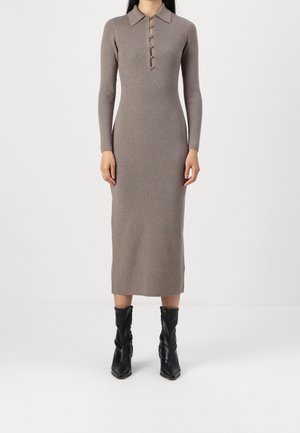 Kobieta ubrana w dopasowaną, długorękawową, prążkowaną sukienkę midi w kolorze taupe z kołnierzem i guzikami z przodu, w połączeniu z czarnymi skórzanymi butami do połowy łydki.