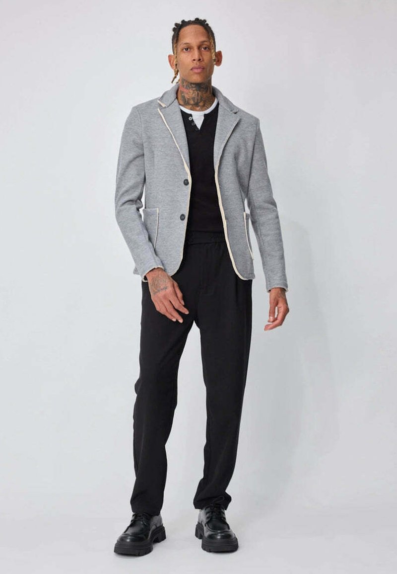 Blazer gris avec un bord crème contrastant, devant boutonné, deux poches, associé à un haut noir et un pantalon noir, porté avec des chaussures noires.