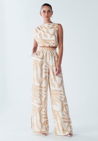 Combinaison large beige avec des motifs abstraits blancs. Fabriquée en tissu léger, avec un haut court et une taille élastique.