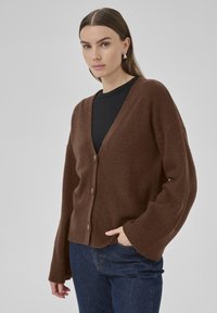 Brauner gestrickter Cardigan mit tiefem V-Ausschnitt, drei Knöpfen und gerippten Ärmeln. Weiche Textur; lässige, lockere Passform. Über einem schwarzen Oberteil getragen.