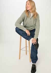 Vaaleanvihreä neulepaita, jossa on puolivetoketju kauluksessa, yhdistetty tummaan sinisiin kapeisiin farkkuihin ja mustiin liukuvaan kengiin. Taustalla puujakkara.