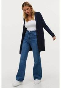 Marineblauer langer Cardigan mit Knopfleiste und Taschen, getragen �über einem weißen Oberteil und hochtaillierten blauen Jeans, kombiniert mit weißen Sneakers.