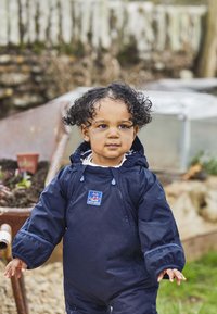 Costum impermeabil de tip salopetă, de culoare bleumarin, cu glugă, dotat cu mansete elastice și o etichetă cu logo. Copilul stă lângă o roabă plină cu pământ.