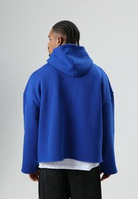 Blauwe oversized capuchontrui van zacht materiaal, met een kangoeroezak en verzakte schouders, gedragen over een wit shirt.