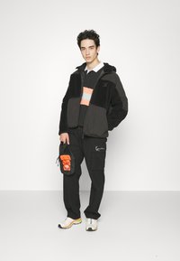 Veste en polaire noire et grise avec fermeture éclair à l'avant, superposée sur une chemise noire avec des rayures orange et grises, assortie à un pantalon cargo et des baskets.