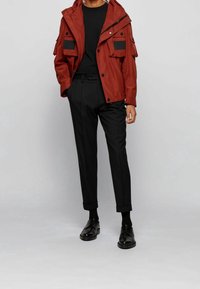 Rote wasserdichte Jacke mit zwei Fronttaschen, kombiniert mit schwarzen maßgeschneiderten Hosen und schwarzen Schuhen. Das Outfit hat ein minimalistisches Design.