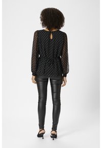 Blouse noire à pois blancs, manches en voile transparent et détail dos avec ouverture en forme de goutte, associée à un pantalon en cuir noir ajusté et des talons noirs.