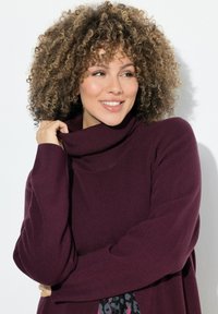 Burgunderfarbener Strickpullover mit hohem Kragen, der eine lockere Passform und gerippten Textur bietet. Die Hände des Models sind vorne im Schoß gefaltet.
