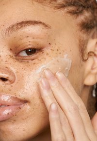 En klar hudvårdsprodukt applicerad på ett ansikte med en varm, mjuk hy och spridda fräknar. Håret är stylat i lösa lockar.