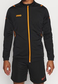 Svart idrottsjacka med orange detaljer och dragkedja. Har en hög krage och långa ärmar. Bärs med svarta shorts och minimal märkning.