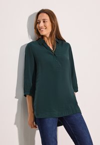 Camicia button-up verde scuro a maniche lunghe con colletto e vestibilità dritta. Presenta un orlo asimmetrico e un tessuto testurizzato. Indossata con jeans blu.