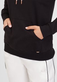 Sudadera negra con capucha, bolsillo tipo canguro, puños y dobladillo acanalados. Presenta cremalleras en tono cobre. Combinada con pantalones blancos con rayas laterales negras.