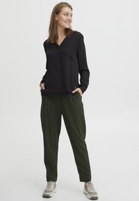 Svart langermet skjorte med brystlomme, kombinert med olivengrønne smale bukser. Modellen har på seg lysebeige joggesko. Enkelt design.