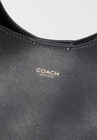 Bolso de cuero negro con costuras plateadas visibles y el logo dorado "COACH NEW YORK" embossado en la superficie.