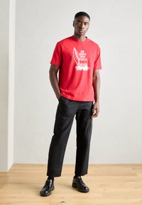 Red cotton t-shirt with white graphic design on front, paired with black cargo pants. Model wears black shoes. Simple, casual outfit.