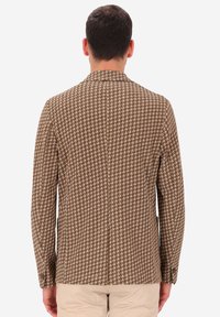 Uomo in blazer a trama houndstooth marrone e pantaloni beige, di spalle su uno sfondo bianco semplice.