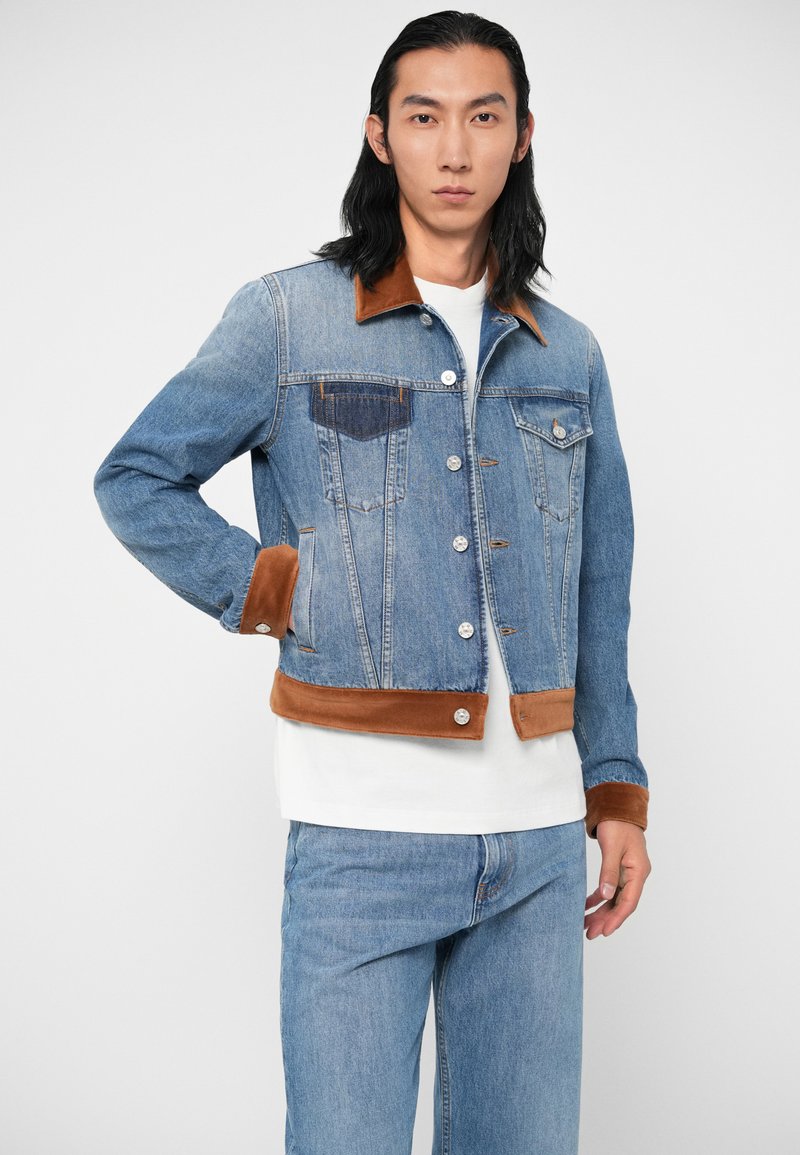 Chaqueta de mezclilla con cuello y puños de pana marrón, cierre de botones, dos bolsillos en el pecho y un diseño ajustado. Color azul claro con una textura suave.