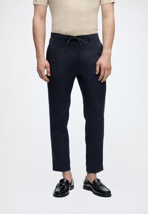 Man draagt marineblauwe broek met trekkoorden en korte pijpen en zwarte leren loafers, staand tegen een effen lichte achtergrond.