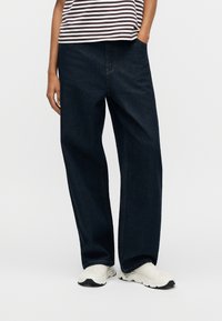 Άτομο που φοράει σκούρο, φαρδύ τζιν, λευκά αθλητικά παπούτσια και ριγέ πουκάμισο, με το ένα χέρι στην τσέπη, στέκεται σε λευκό φόντο.