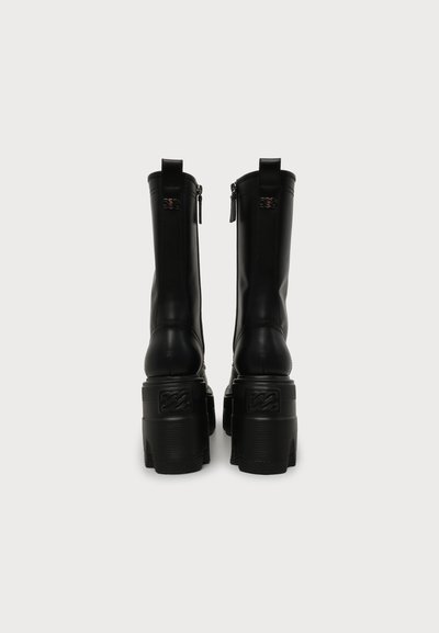 Paire de bottes hautes en cuir noir avec des talons hauts épais et des fermetures éclair à l'arrière, présentées de dos sur un fond clair uni.