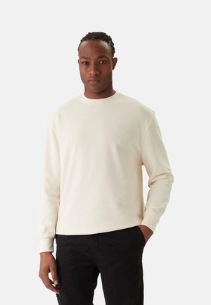 Hombre con cabello trenzado, vestido con un suéter de manga larga color crema y pantalones negros, de pie con una mano en el bolsillo contra un fondo liso.