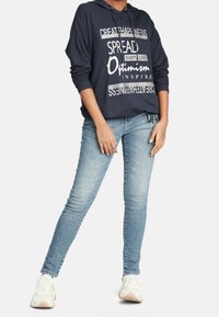 Marinblå hoodie med ett framstående grafiskt motiv i vitt och silver, kombinerad med ljusblå skinny jeans och vita sneakers med beige detaljer.