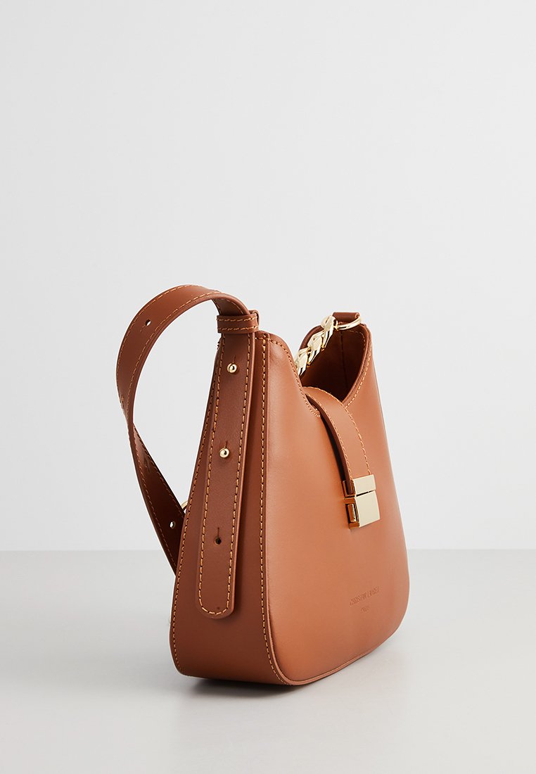Sac à main en cuir marron avec bandoulière réglable, fermoir doré et détail de chaîne, sur fond clair uni.