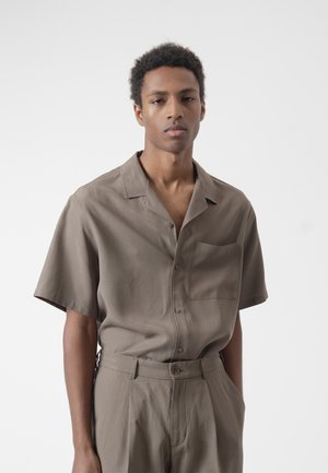 Joven con cabello corto y rizado, vestido con una camisa taupe de manga corta con botones y pantalones de cintura alta a juego sobre un fondo blanco liso.