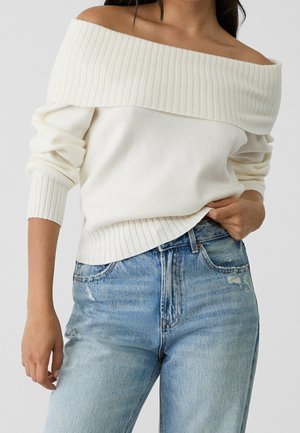 Woman wearing off-shoulder white ribbed sweater and light blue high-waisted jeans with slight distressing on pocket.