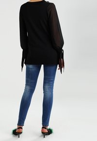 Schwarzes Langarmoberteil mit transparenten Ärmeln und gebundenen Manschetten, kombiniert mit figurbetonten blauen Jeans und schwarzen High Heels mit grünen Fellakzenten.