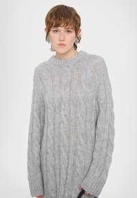 Pulover gri deschis, tricotat cu model cable, cu guler rotund, croială lejeră și detalii cu textură. Mâneci lungi cu manșete ribbata. Material moale, transparent.