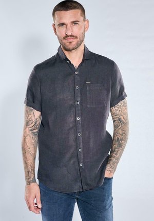 Mann mit kurzen Haaren und Tattoos auf beiden Armen, trägt ein kurzärmeliges schwarzes Hemd mit Knöpfen und blaue Jeans, steht vor einem einfarbigen Hintergrund.