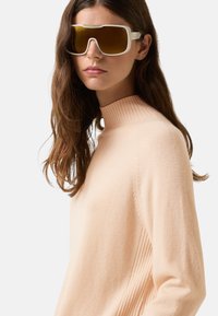 Vrouw in een oversized witte bril met bruin getinte glazen en een lichtbeige geribde coltrui, iets naar rechts gekeerd.