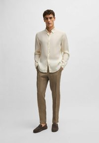 Camisa button-up color beige claro con cuello clásico, combinada con pantalones de lino marrón y zapatos oscuros de tipo slip-on. Textura de tela suave.