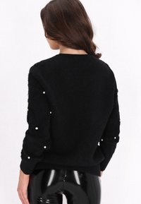 Schwarzer Strickpullover mit strukturierten Ärmeln und Perlenakzenten. Figurbetonter Schnitt, runder Halsausschnitt und gerippte Manschetten. Kombiniert mit glänzenden schwarzen Hosen.