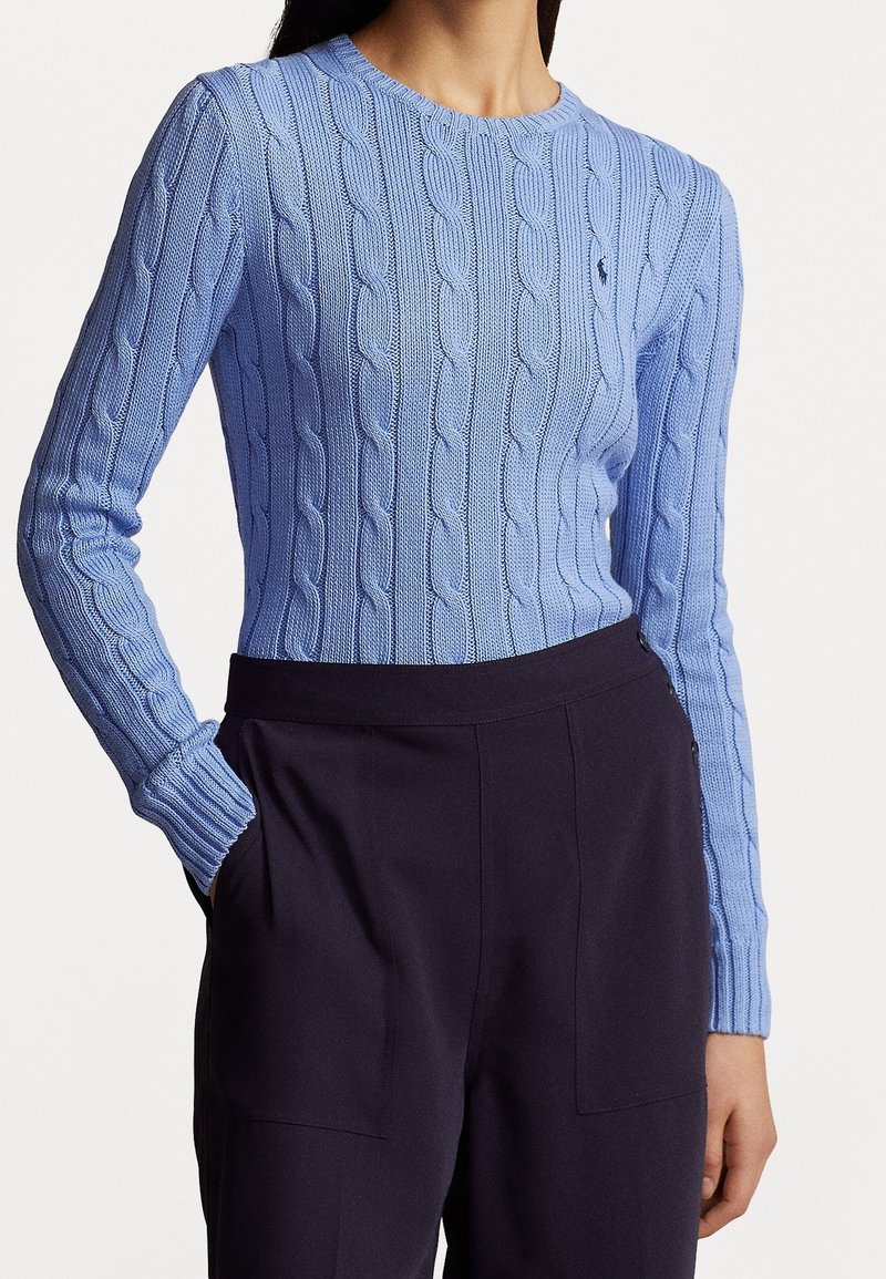 Personne portant un pull bleu clair torsadé avec un petit logo sombre sur la poitrine, associé à un pantalon noir taille haute.