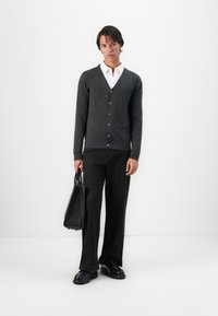 Giovane uomo in piedi che indossa un cardigan grigio scuro sopra una camicia bianca, pantaloni neri, scarpe nere, mentre tiene una borsa in pelle nera.