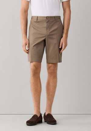 Mann trägt beige knielange Shorts, weißes Hemd und dunkelbraune Loafers, steht vor einem schlichten grauen Hintergrund.