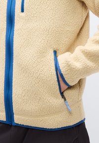 Μπεζ φλις μπουφάν με μπλε φερμουάρ και περιγράμματα, χέρι μερικώς μέσα στην φερμουάρ τσέπη στο πλάι, φορεμένο πάνω από μαύρο παντελόνι.