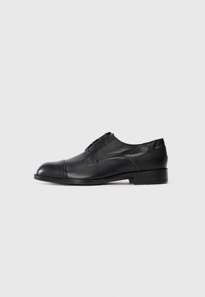 Chaussure Oxford homme en cuir noir avec bout rapporté, fermeture à lacets et talon bas sur fond gris clair.