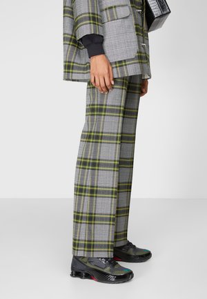 Osoba nosząca szare i żółte spodnie w kratę z dopasowaną marynarką oraz czarne i zielone buty sportowe, trzymająca czarną teksturowaną torebkę.